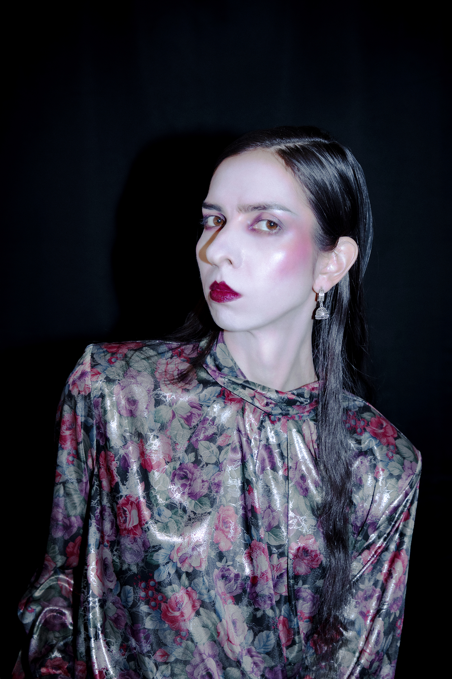 Female model with pale glassy skin and deep blush and lip in a reflective blouse looking towards the camera.